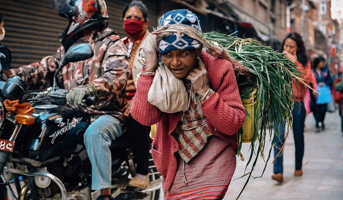 Gender Gap Nepal, woman carries a bundle of vegetables on a busy urban street filled with people and motorcycle